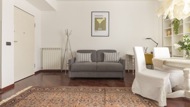 Un salon avec un canapé gris, un tapis à motifs, un tableau encadré au mur et une chaise blanche.