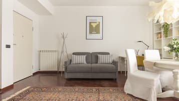 Un soggiorno con un divano grigio, un tappeto decorato, un quadro appeso al muro e una sedia bianca.