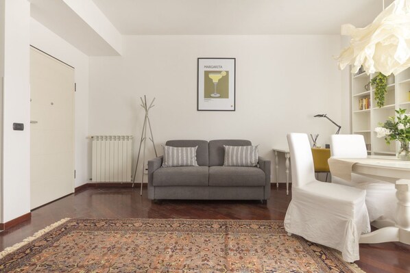 Un salon avec un canapé gris, un tapis à motifs, un tableau encadré au mur et une chaise blanche.