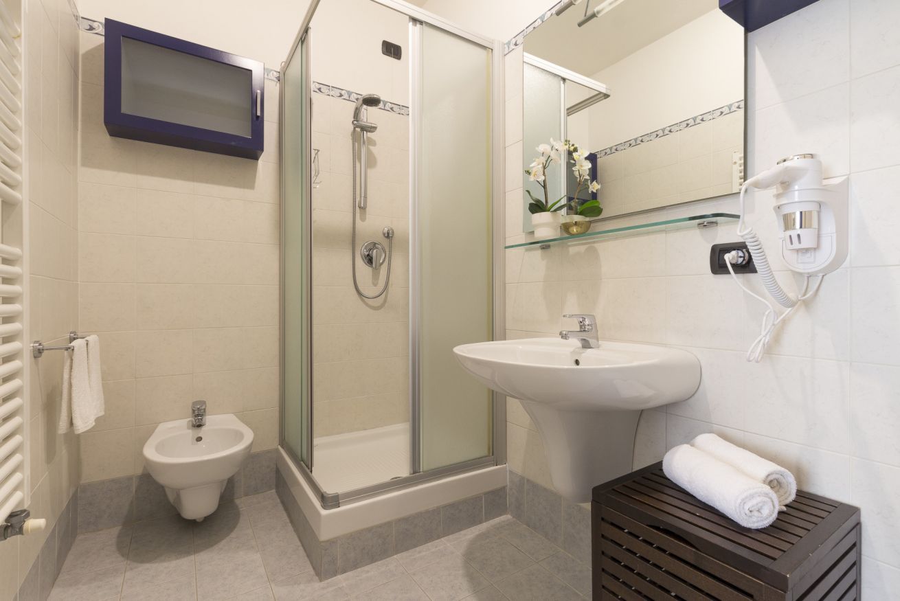 Et badeværelse med glasvæg omkring bruseniche, et konsolbord med vask, en bidet og hårtørrer samt et håndklædestativ.