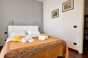 Una cama bien hecha con sábanas blancas y una colcha estampada, una sola almohada amarilla y dos cuadros en la pared.