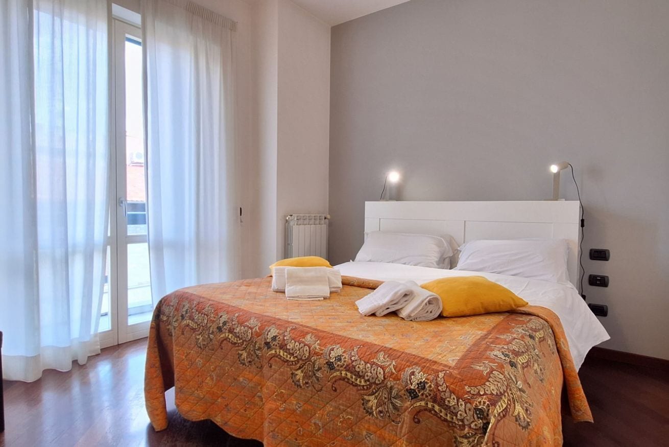 A hotel room with a bed, a radiator, a window with curtains, and a wall-mounted light.