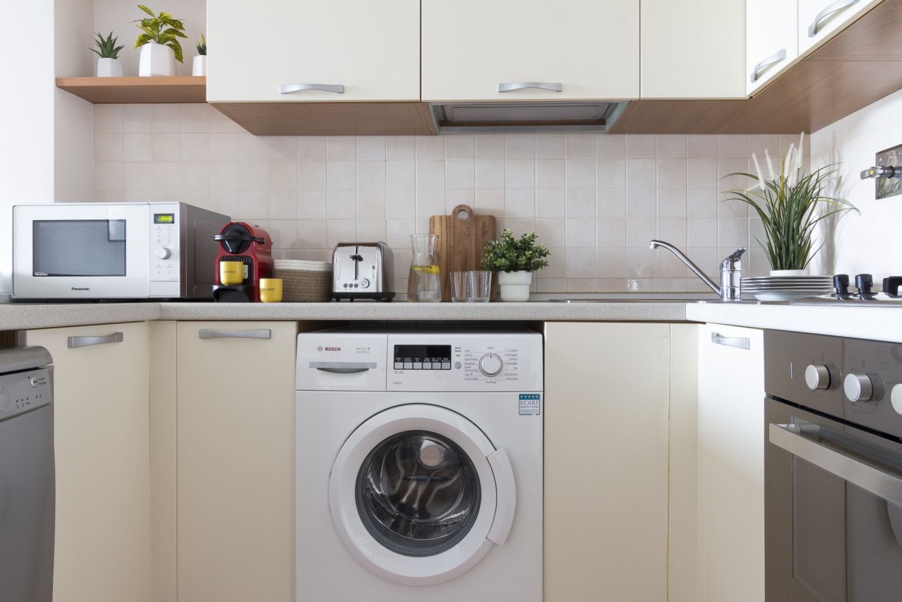 A compact kitchen with a washing machine, microwave, toaster, and a small countertop with potted plants.