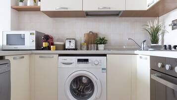 A compact kitchen with a washing machine, microwave, toaster, and a small countertop with potted plants.