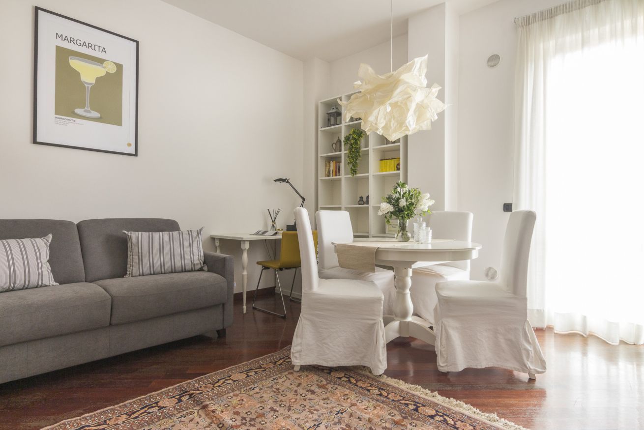 Ein Wohnzimmer mit grauem Sofa, einem Esstisch mit weißen Stühlen, einem Bücherregal und einer großen dekorativen Pendelleuchte.