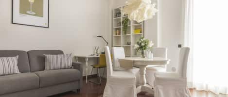 Ein Wohnzimmer mit grauem Sofa, einem Esstisch mit weißen Stühlen, einem Bücherregal und einer großen dekorativen Pendelleuchte.
