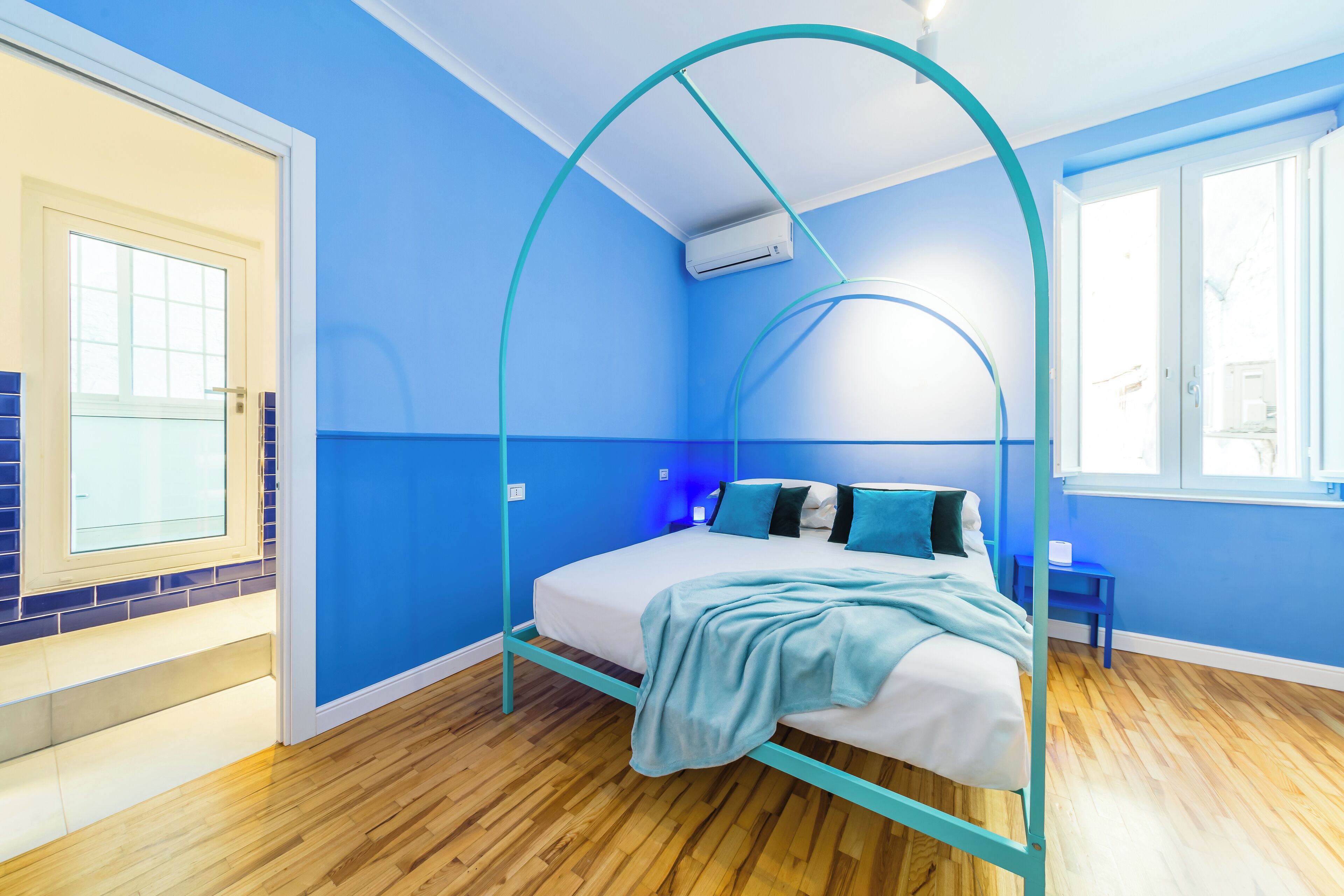 A bedroom with a bed under a blue canopy, a window, and a wooden floor.