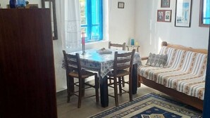 Un coin repas avec une table ronde et des chaises, un canapé agrémenté de coussins à motifs et une fenêtre bleue avec des rideaux blancs.