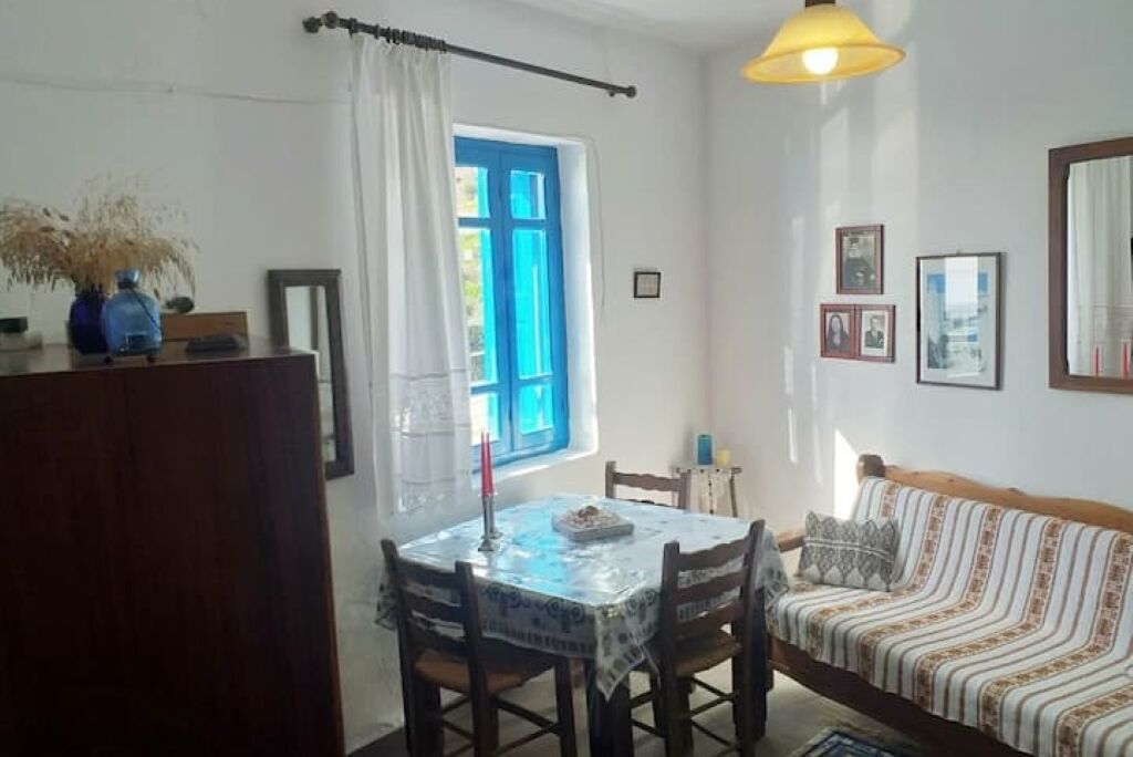 A dining area with a table and chairs, a sofa, a window with blue shutters, and a hanging lamp.