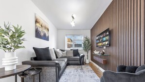 A modern living room with a sectional sofa, a coffee table, and a TV mounted on a wooden wall. - Spacious 4BR Home in Brentford, Free Parking (Brentford)