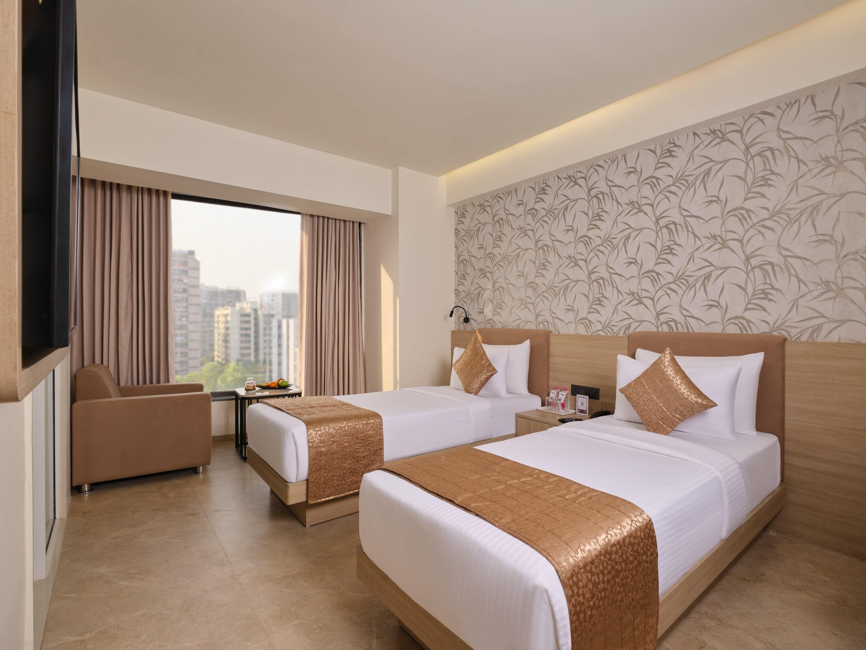 A hotel room with two beds, a TV, a desk, and a view of the city.