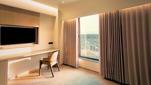 Een moderne hotelkamer met een grote televisie aan de muur, een bureau met stoel, en een groot raam met gordijnen.