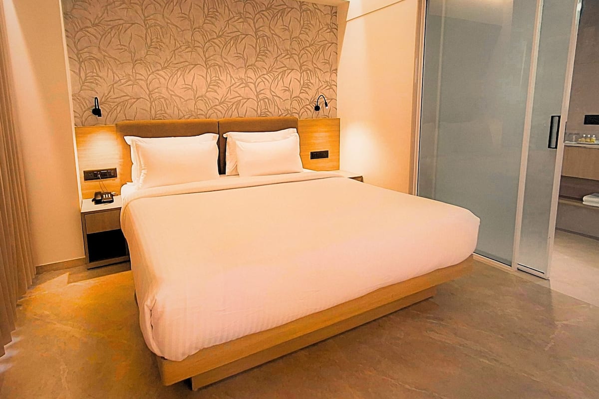 Een moderne hotelkamer met een groot bed, twee nachtkastjes met lampen, een hoofdbord met bladmotief en een inloopkast met glazen schuifdeuren.