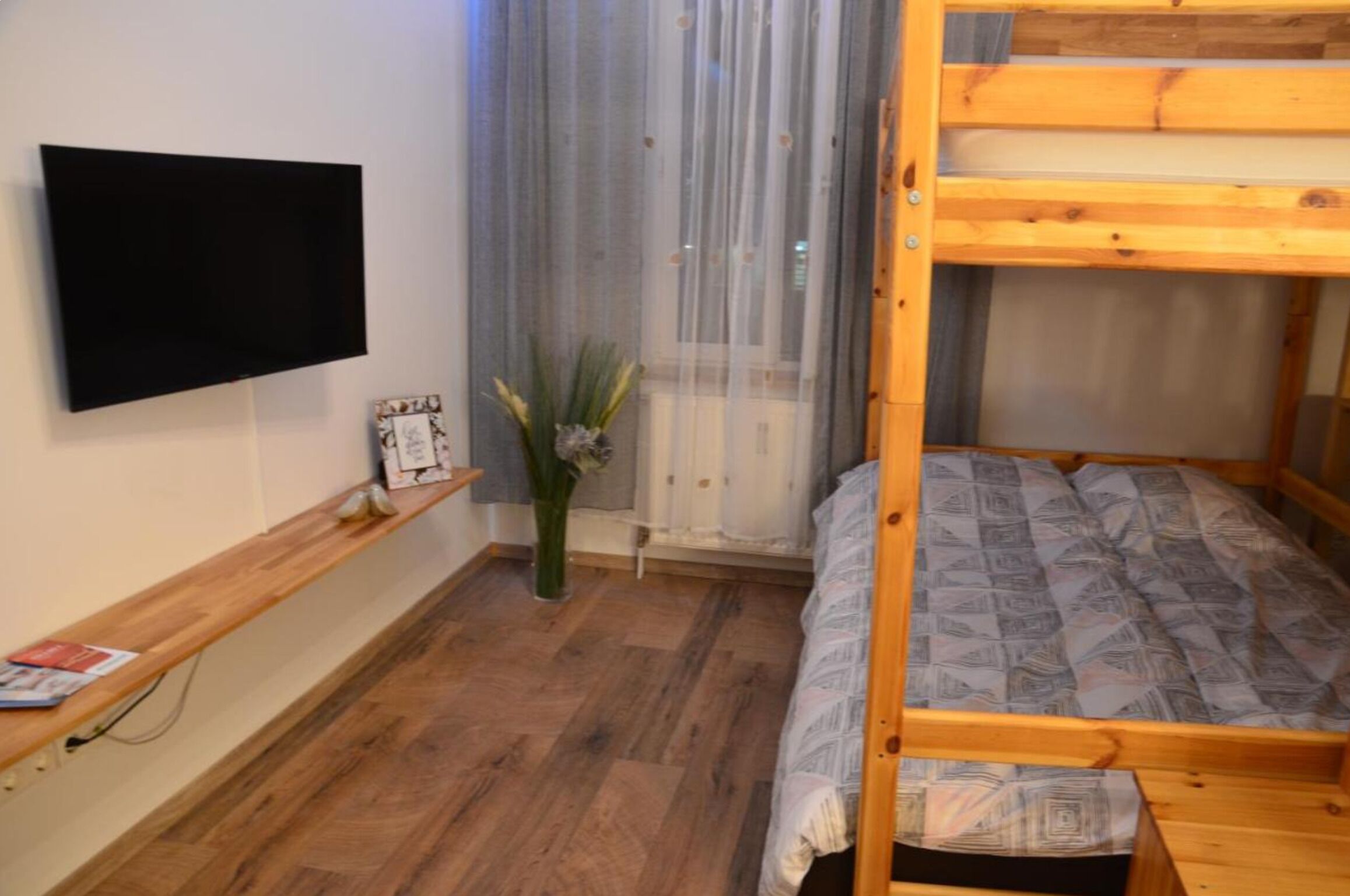 Une chambre avec un lit superposé, un téléviseur à écran plat fixé au mur, une étagère en bois ornée d’un vase et d’une petite plante, et une fenêtre avec des rideaux légers.