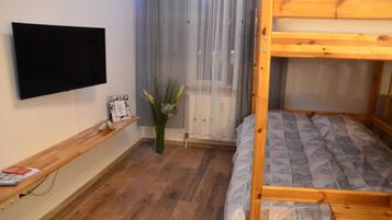 Une chambre avec un lit superposé, un téléviseur à écran plat fixé au mur, une étagère en bois ornée d’un vase et d’une petite plante, et une fenêtre avec des rideaux légers.