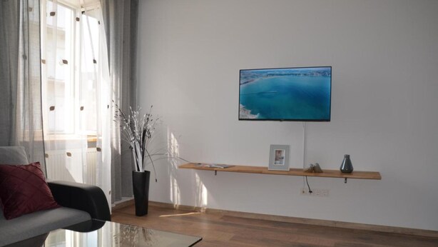 A modern living room with a large flat-screen TV mounted on the wall, a glass coffee table, a sofa, and a wooden shelf with decorative items. - Vega - Free Parking - Self Checkin (Graz)