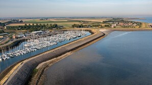 Eine Luftaufnahme eines Hafens mit zahlreichen Booten, die an einer Uferpromenade vertäut sind, umgeben von Feldern und einer kleinen Stadt im Hintergrund.