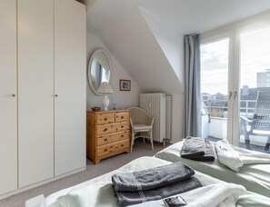 A bedroom with a bed, a dresser, a chair, a radiator, and a window with a view of buildings. - Ferienwohnung in Busum (Büsum)