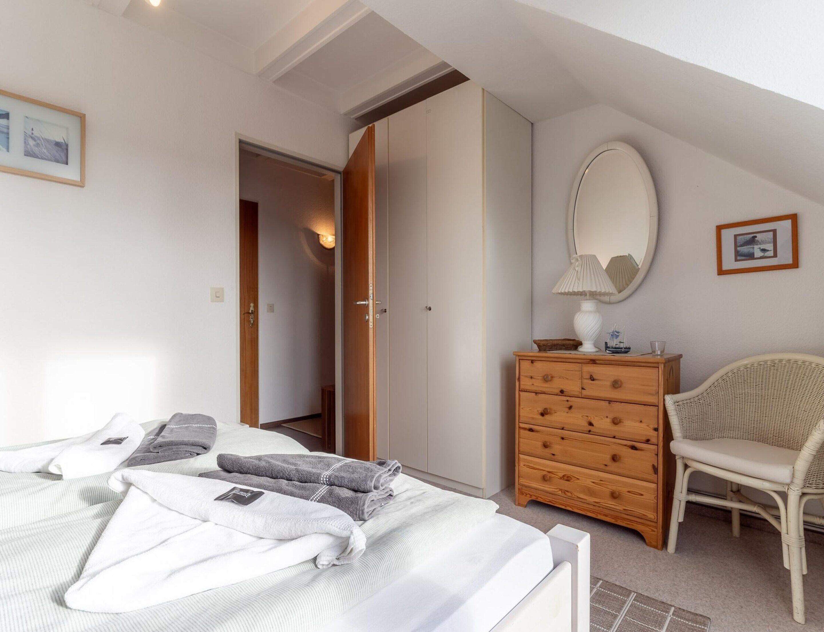 Une chambre à coucher comprenant un lit, une commode en bois, une chaise en osier, un miroir et une porte donnant sur une autre pièce.