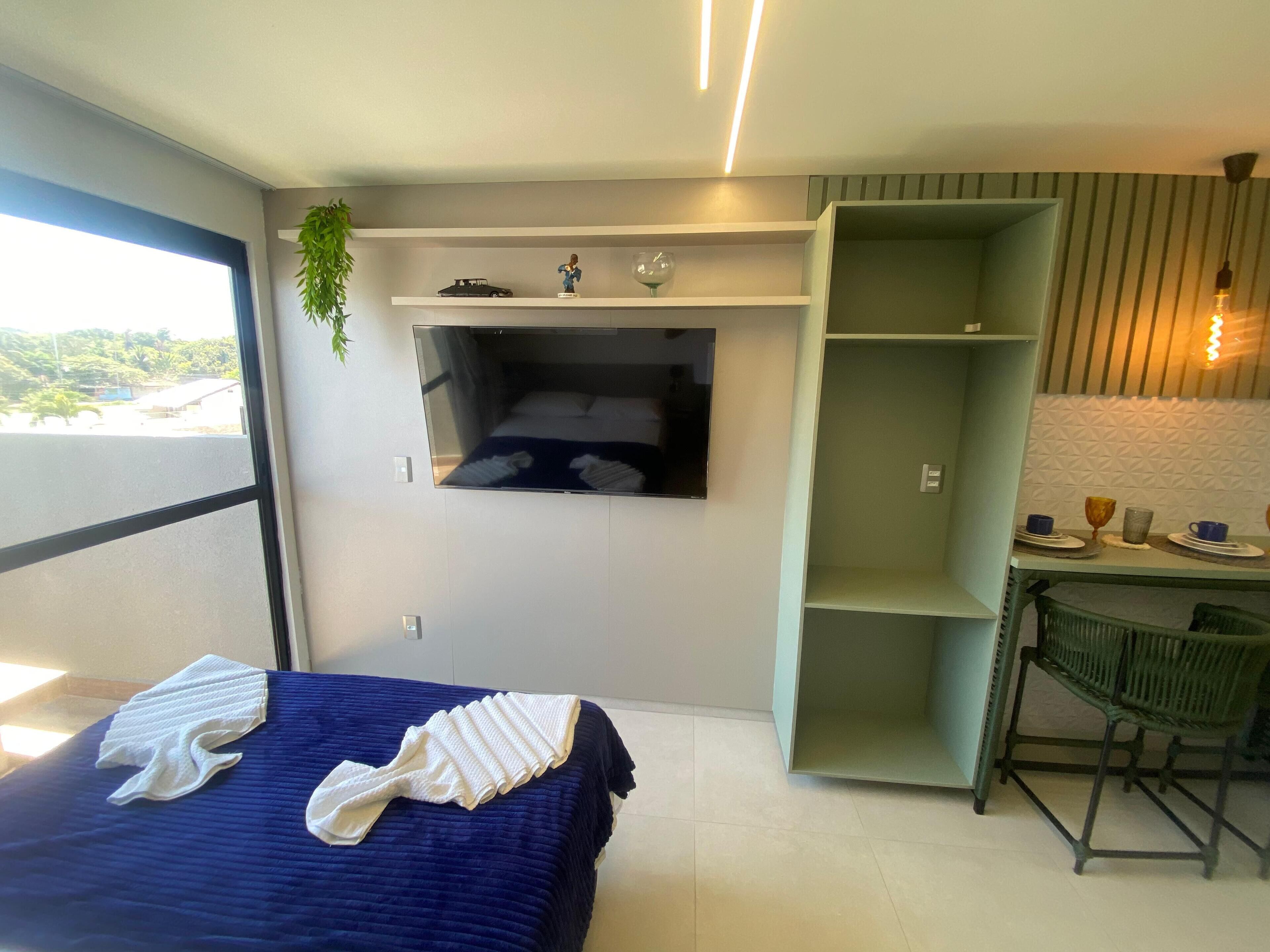A modern hotel room with a bed, a TV mounted on the wall, a small table with cups and a glass, and a green chair.