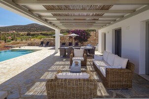 Una zona de estar al aire libre techada, con suelo de piedra, mobiliario de mimbre y piscina.