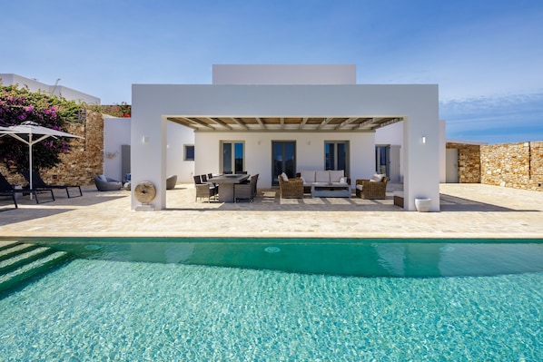 Un espace piscine extérieur moderne, avec un bâtiment blanc, du mobilier d’extérieur et un mur en pierre.