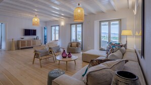 Amplia sala de estar con piso de madera, un sofá grande, una mesa de centro redonda y un televisor montado en la pared. Hay dos lámparas colgantes, una ventana con vistas y una puerta que da a otra habitación.