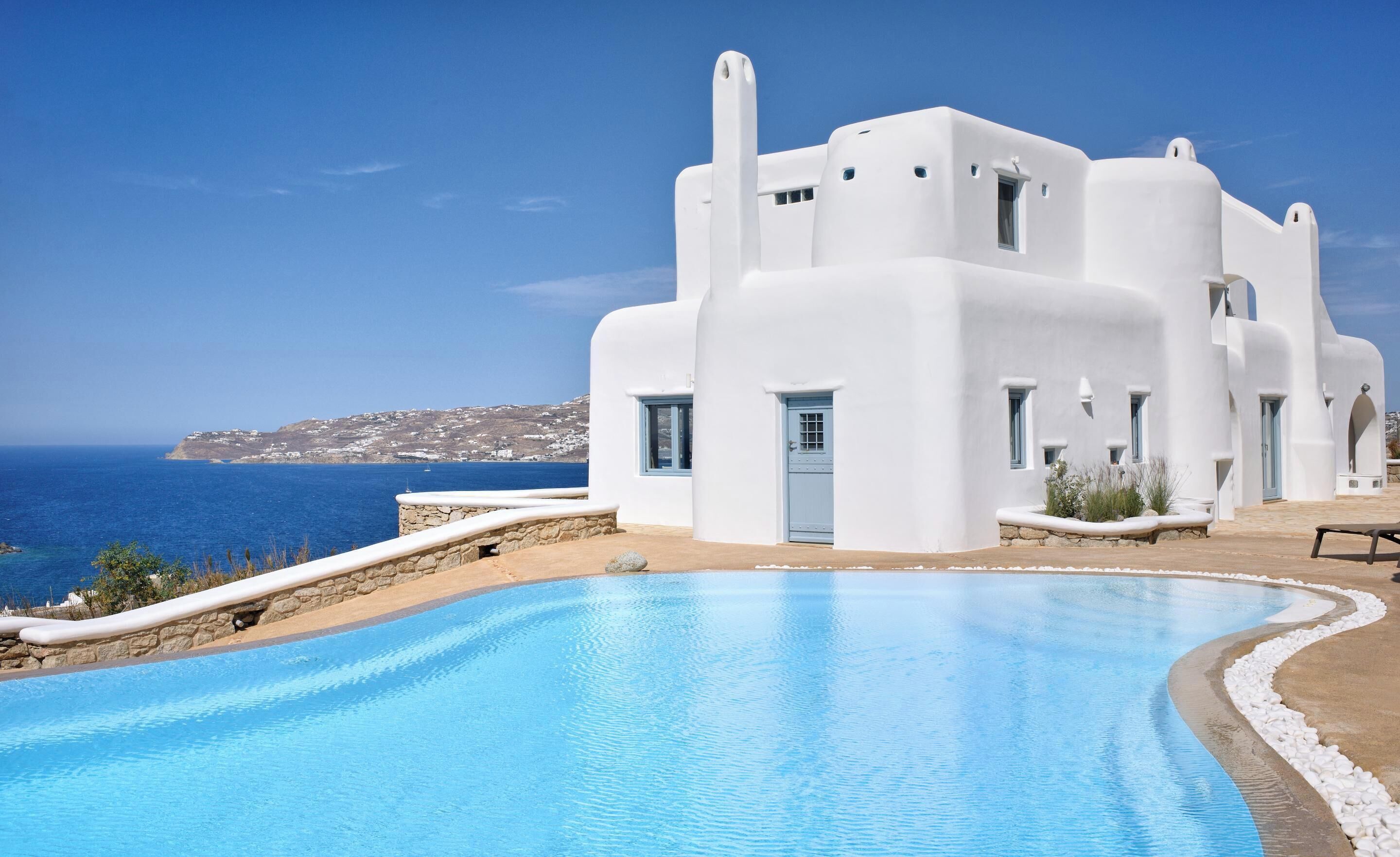 A white building with a pool, a view of the sea, and a stone wall.