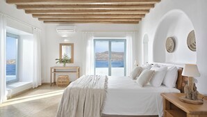 Une chambre avec un grand lit, une tête de lit en bois, une table de chevet avec une lampe, et une vue sur la mer grâce à de grandes fenêtres.