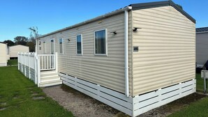 A single-story mobile home with a flat roof, white exterior, and a small porch. - 3 Bedroom Caravan - Pets, Parking, Pool Access (Cirencester)