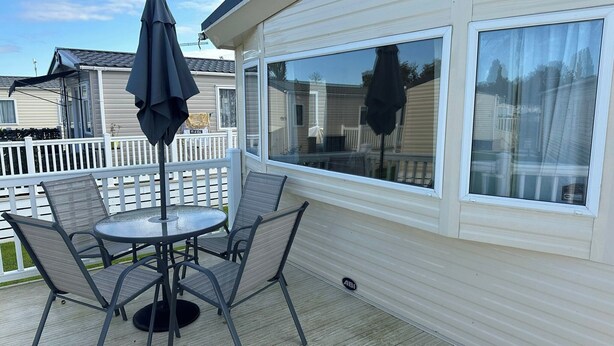 A covered patio with a table and chairs, a large window, and a white fence in the background. - 3 Bedroom Caravan - Pets, Parking, Pool Access (Cirencester)