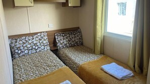 A compact room with two beds, each with floral-patterned bedding and a brown headboard. There is a small shelf above the beds with a lamp and a small container. A window with curtains is visible, and a towel is placed on the floor near the window. - 3 Bedroom Caravan - Pets, Parking, Pool Access (Cirencester)