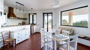 A modern kitchen with white cabinetry, a dining area with a wooden table and white chairs, and a large window offering a view of greenery.