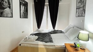 Een slaapkamer met een bed, een nachtkastje met een lamp, een raam met gordijnen en twee ingekaderde foto's aan de muur.