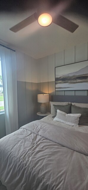 A bedroom with a bed, a nightstand with a lamp, a ceiling fan, and a painting on the wall.