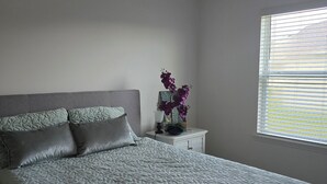 A bedroom with a bed, pillows, a nightstand, a lamp, and a window with blinds. - Home away from Home (Port St. Lucie)