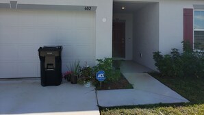 A residential garage with a number 602 garage door, a black trash bin, and a small garden with a blue sign. - Home away from Home (Port St. Lucie)