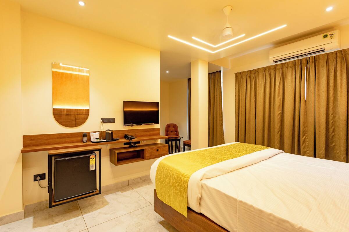 A hotel room with a bed, a TV, a desk with a chair, a small fridge, and a window with curtains.