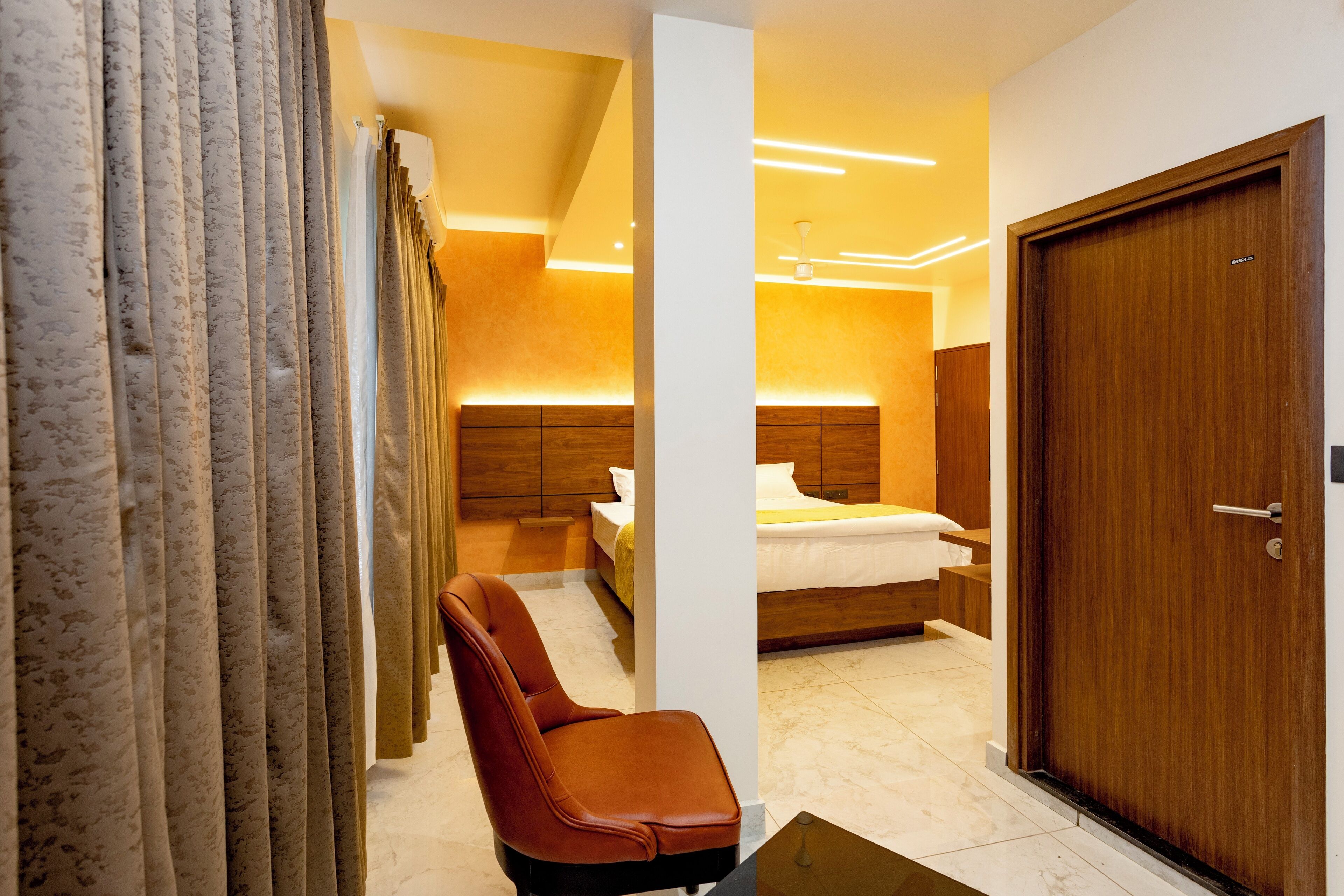 A modern hotel room with a wooden door, a bed with white bedding, a nightstand, a chair, and a large window with curtains.