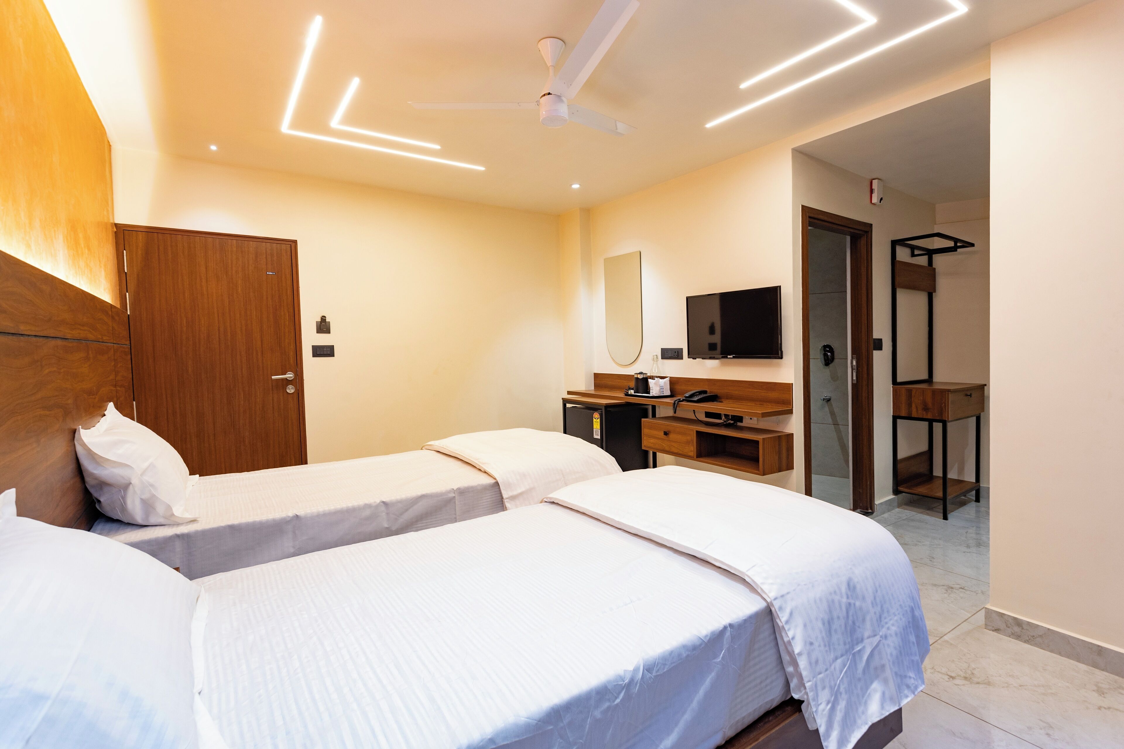 A hotel room with two beds, a wooden wardrobe, a desk with a mirror, and a ceiling fan.