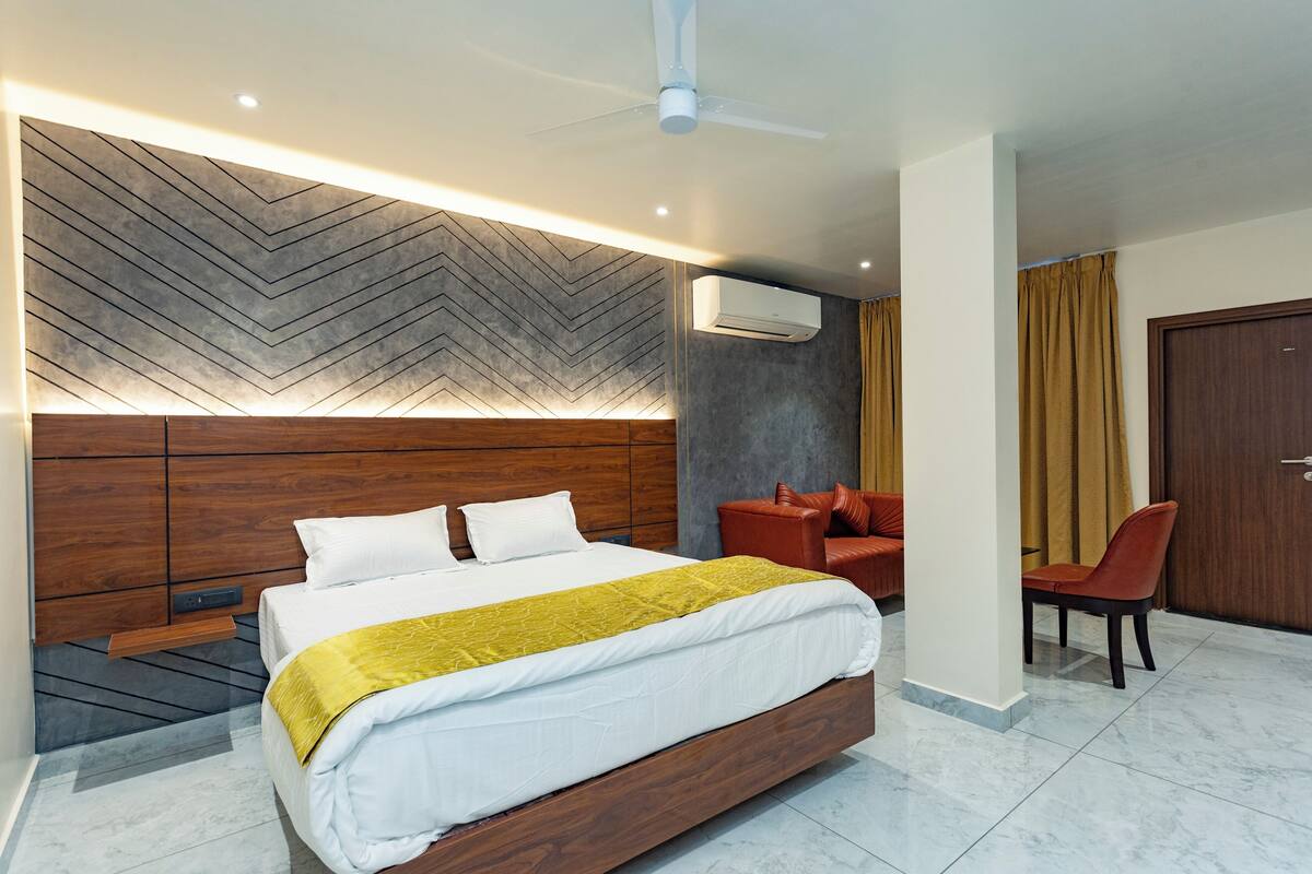 A modern hotel room with a large bed, a wooden headboard, a ceiling fan, an air conditioning unit, a seating area with a red chair, and a door.