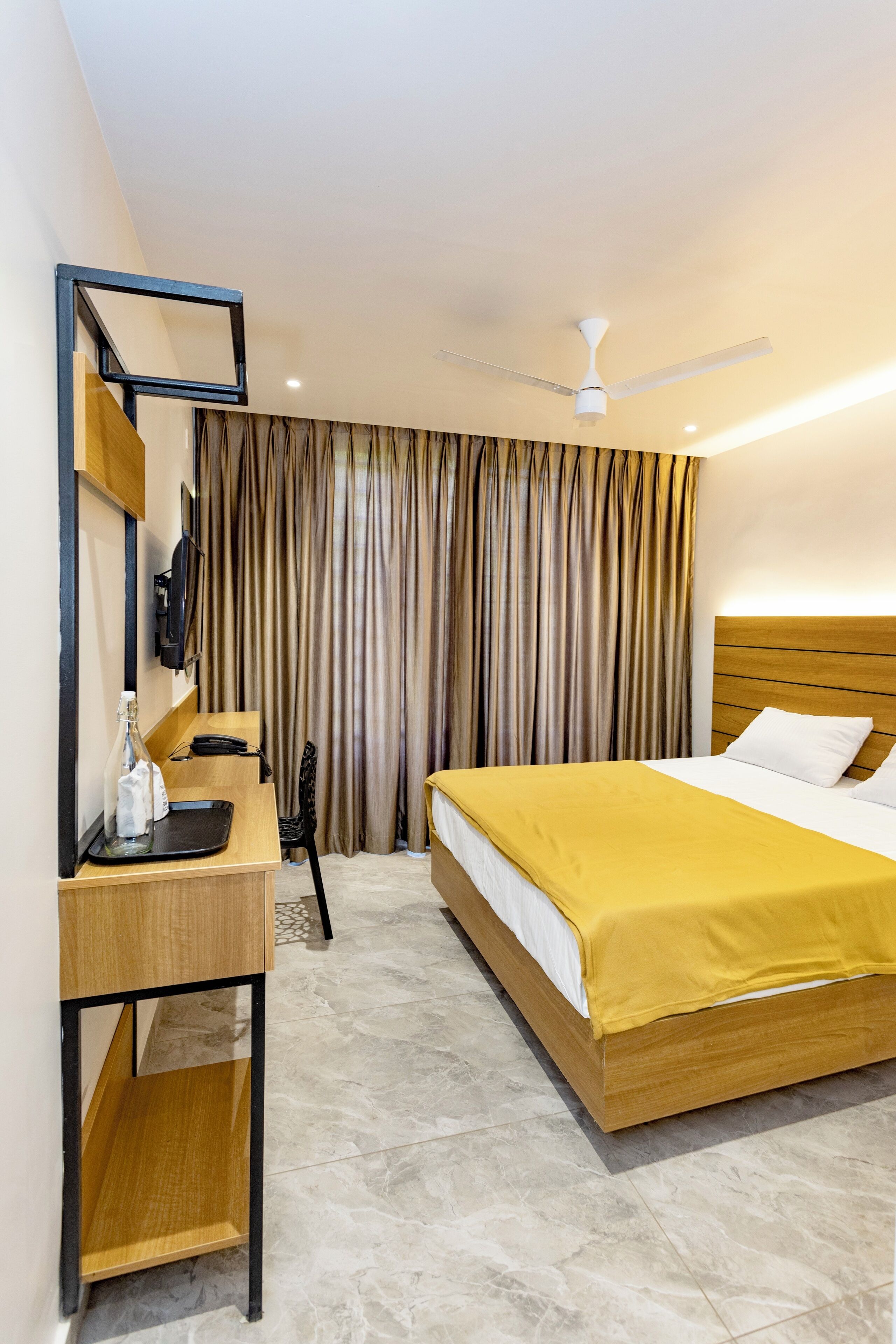 A hotel room with a bed, a desk with a chair, a mirror, a ceiling fan, and a curtain.
