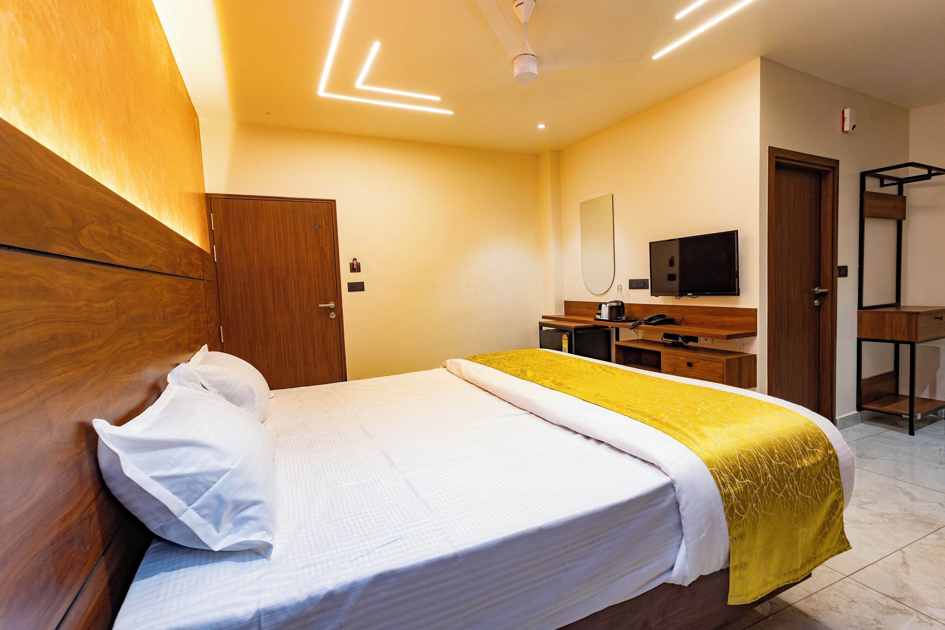 A modern hotel room with a wooden headboard, a bed with white bedding and a yellow throw, a desk with a computer, and a TV.