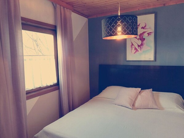Un dormitorio con cama, ventana con cortinas, una obra de arte en la pared y una lámpara colgante.