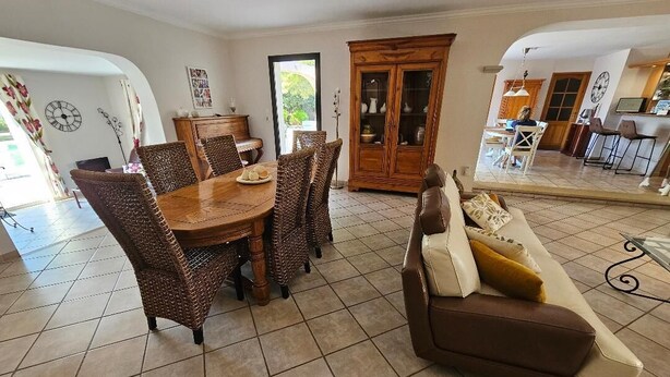Un coin repas avec une table en bois et des chaises en osier, un canapé marron avec des coussins et un meuble avec des portes en verre.