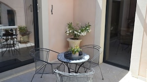 Une terrasse avec une table ronde et des chaises, un pot de fleurs et une porte vitrée donnant sur un espace intérieur.