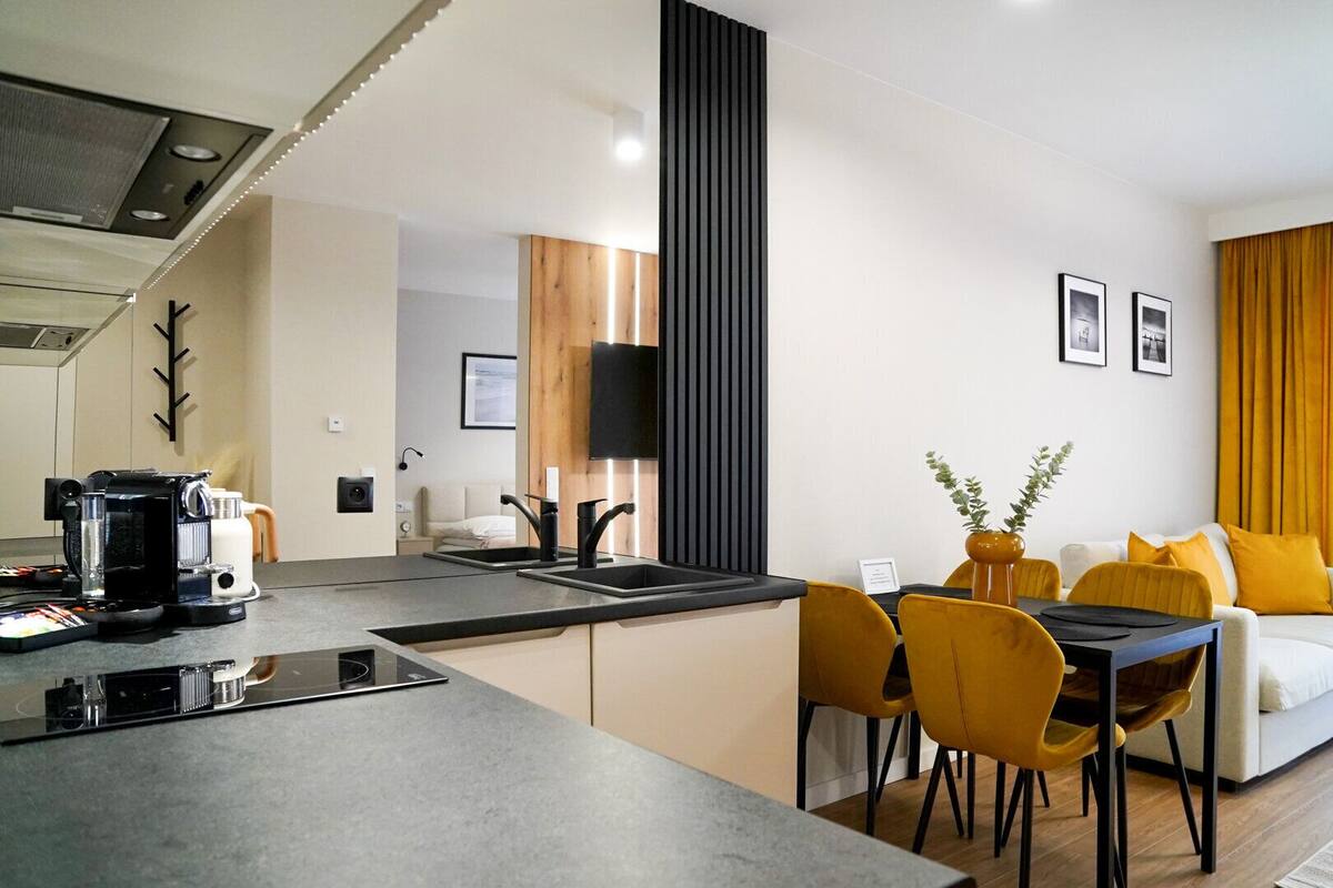 A modern kitchen with a coffee machine, a dining area with yellow chairs, and a living room with a sofa and curtains.