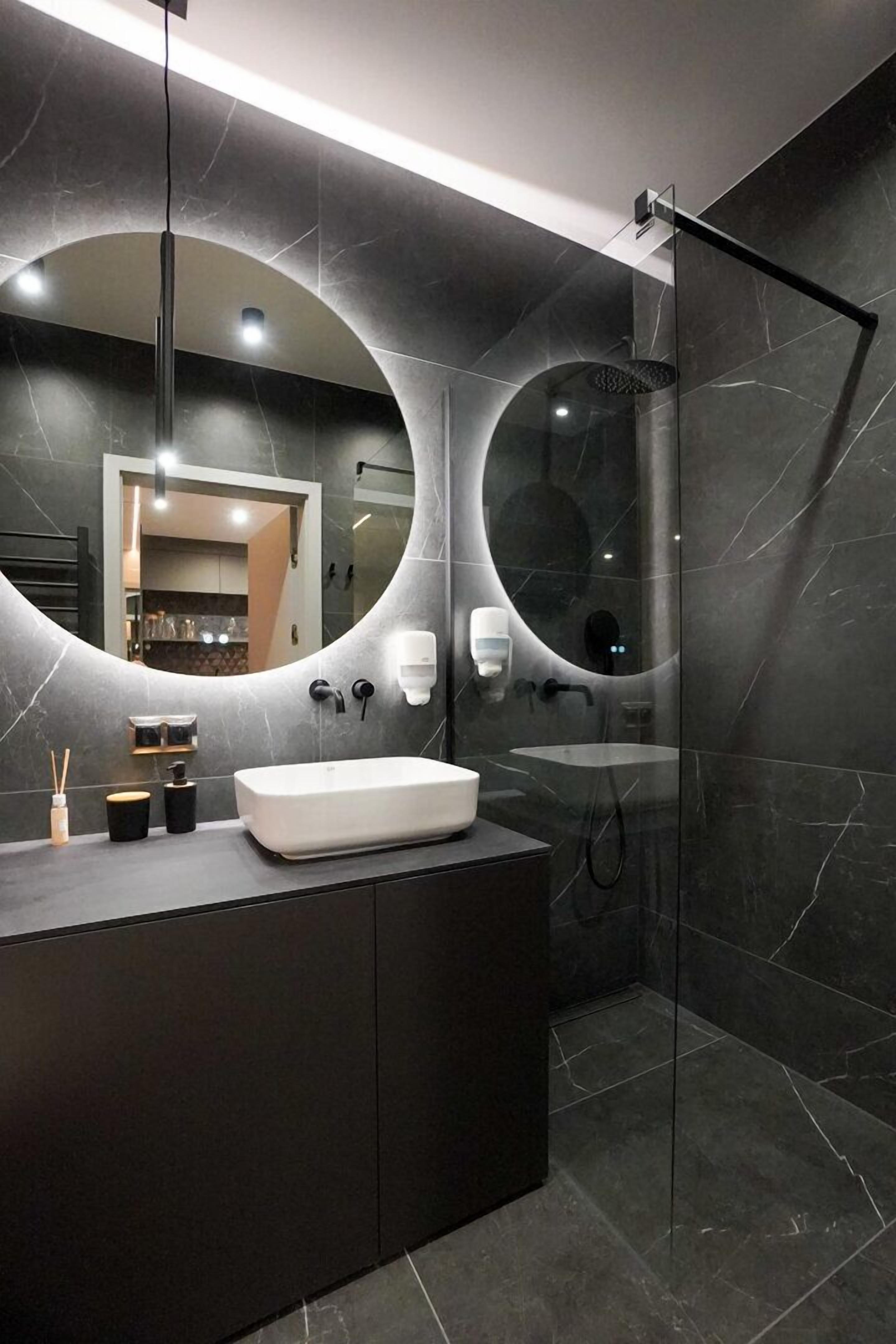 Un baño moderno con un espejo ovalado grande, un lavabo blanco y una ducha con cerramiento de vidrio.