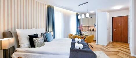 A hotel room with a large bed, a desk, a chair, and a kitchenette in the background.