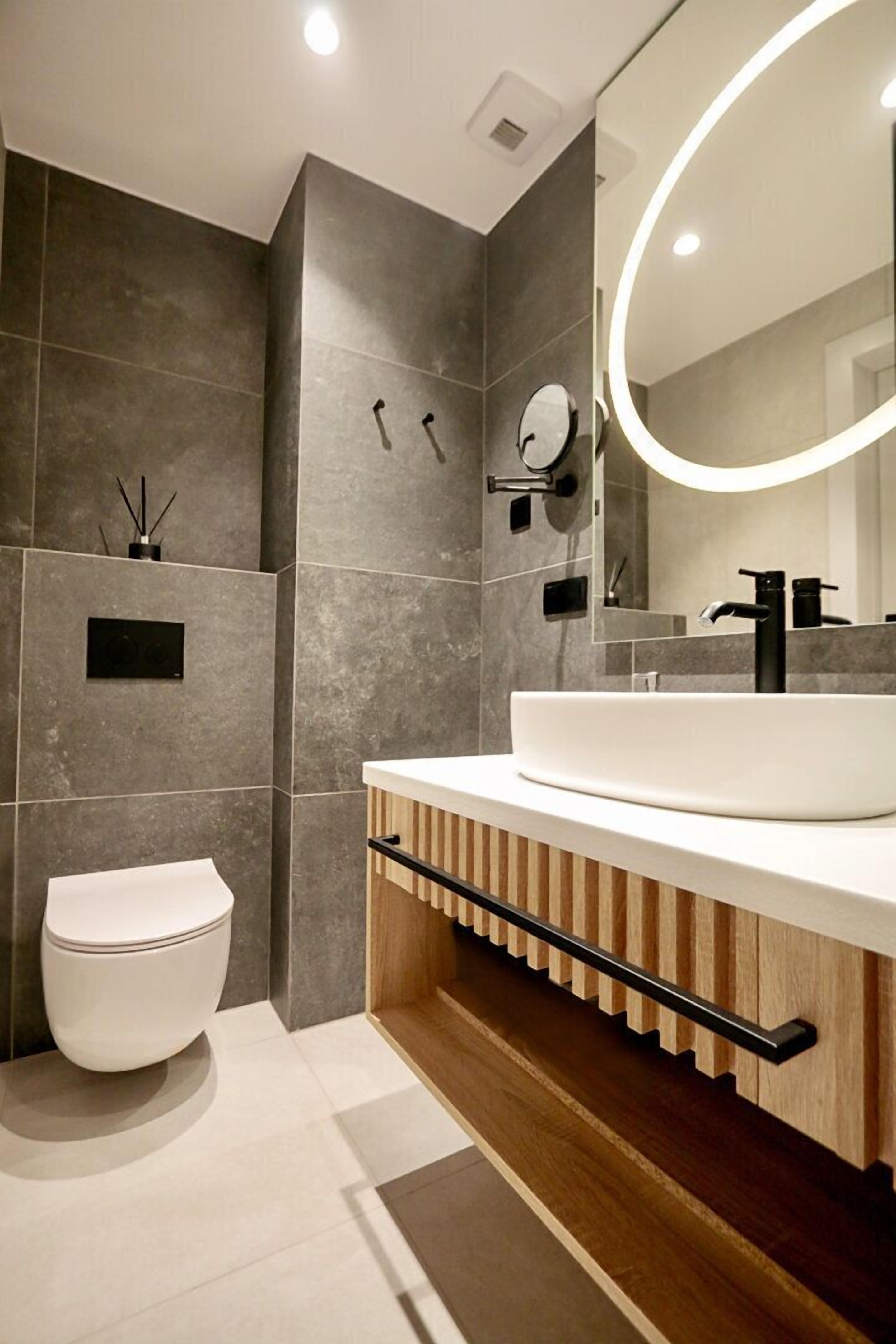 Ein modernes Badezimmer mit einem großen weißen Waschbecken, einem runden Spiegel mit Beleuchtung und einer Toilette.